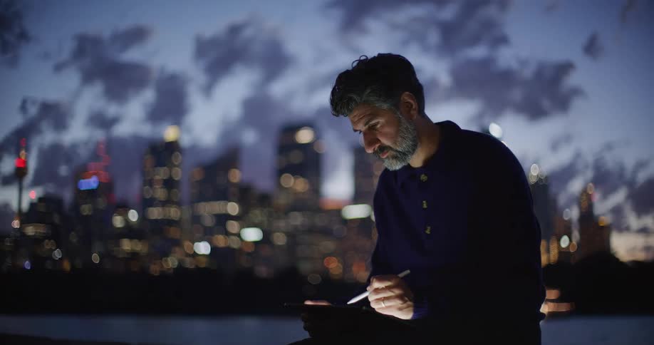 Man uses a digital stylus on a tablet in front of an illuminated cityscape at dusk