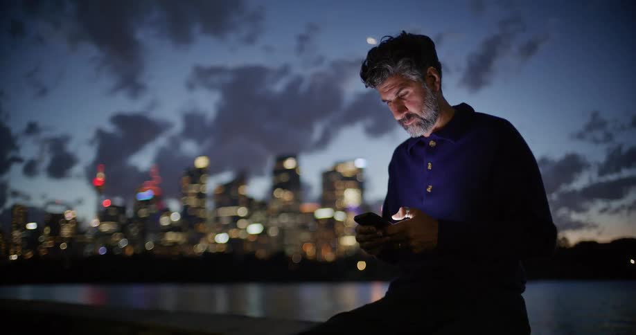 Man sitting along the coast scrolling on his phone as lights from downtown towers and buildings glow in the distance