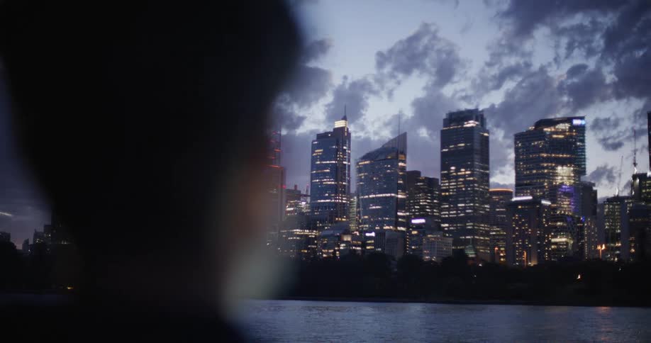 The video shows the sydney skyline at dusk captured from across a body of water with a persons silhouette in the blurred foreground the city lights are illuminated against a deep blue sky filled with textured clouds