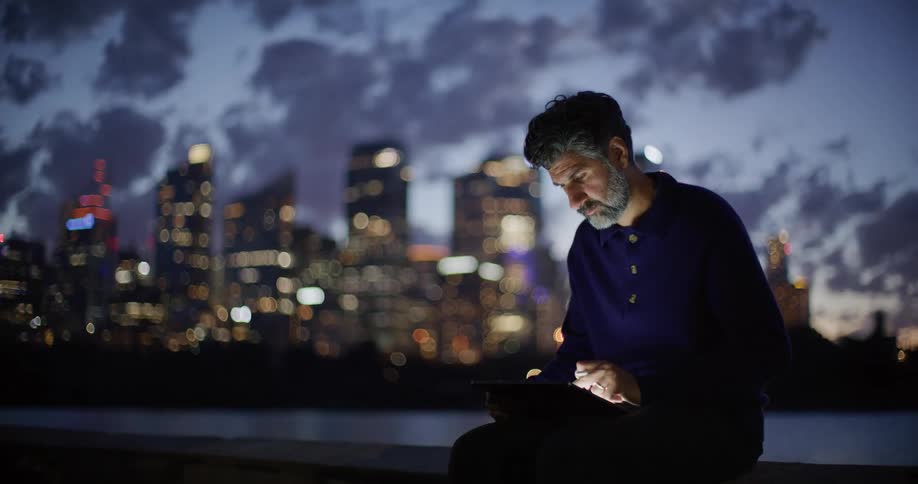 A man uses a stylus on a tablet at night against a backdrop of blurred city lights