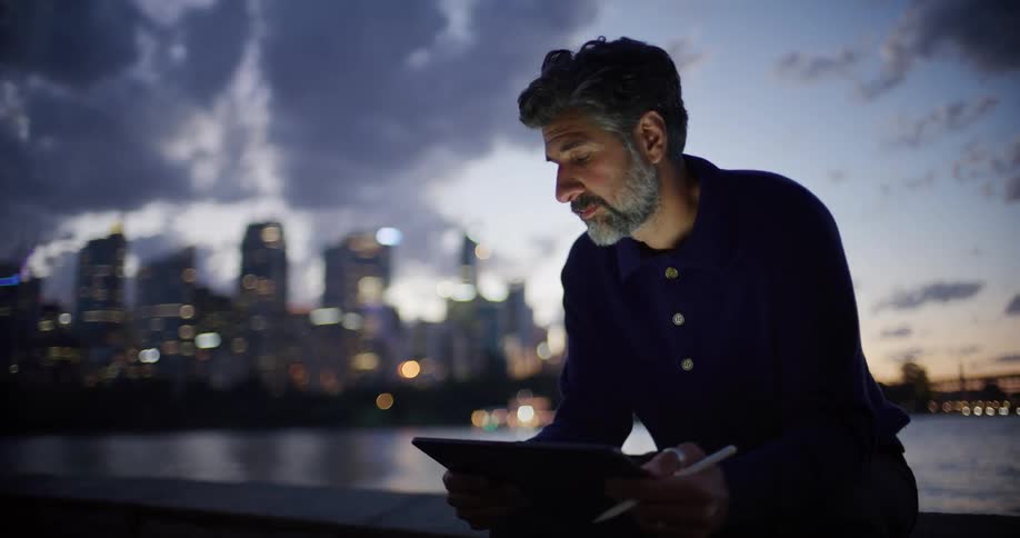 Sitting by the river at dusk a bearded man gazes across at the shimmering city skyline before lowering his eyes to write on his digital tablet with a stylus