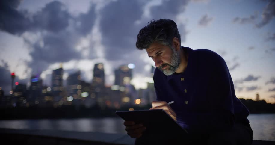 Man writes on a tablet with a digital stylus in front of a city illuminated at dusk