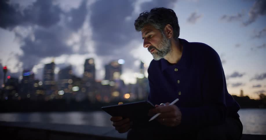 A man with a grey beard sits outdoors at dusk and uses a stylus to draw on a tablet while a city skyline glows in the background