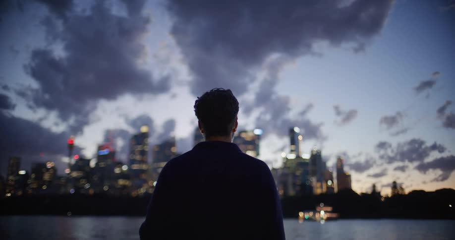 A view from behind a man shows him looking at a bright city skyline across the water at twilight with dark clouds and a blue sky above the illuminated buildings