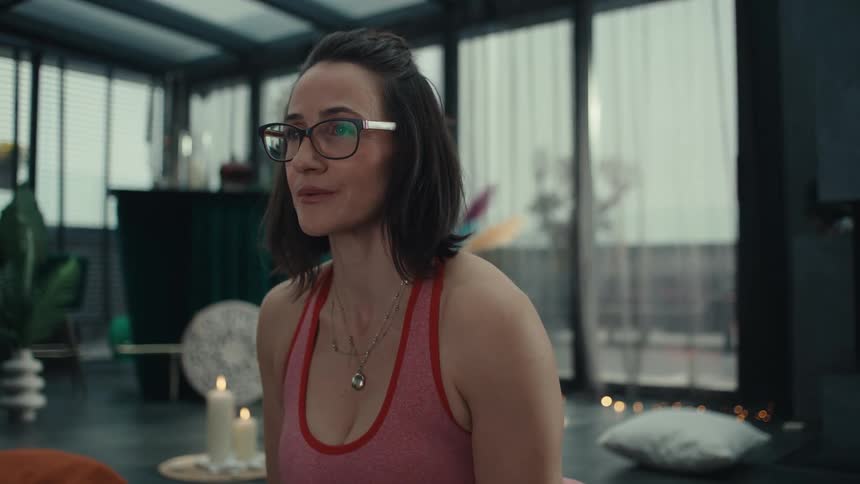 A young woman wearing glasses and pink activewear stretches on a yoga mat indoors at midday