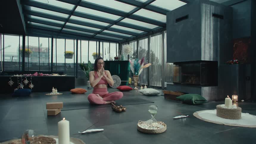 A woman in pink sportswear sits on a yoga mat in a glass walled room raising her arms and bringing her hands to a prayer position before meditating amidst candles cushions and wellness accessories