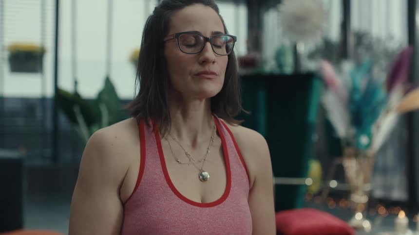 A woman wearing glasses and pink athletic wear sits cross legged on a yoga mat lifts a singing bowl and closes her eyes in a meditative pose