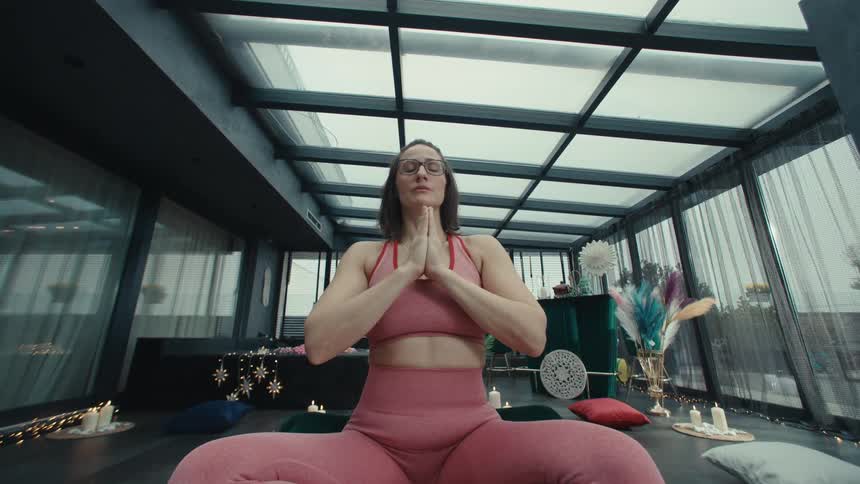 Woman wearing glasses sits on a couch enjoying a warm cup of tea and talking to a companion in the second video the same woman practices yoga under a glass ceiling moving from an overhead stretch into a seated meditative pose