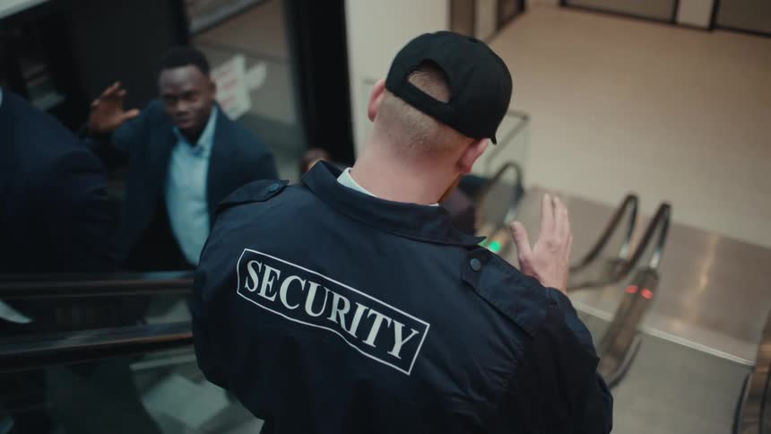 A view from behind a security guard as he descends an escalator into a busy modern building lobby where several people in business attire are walking and talking