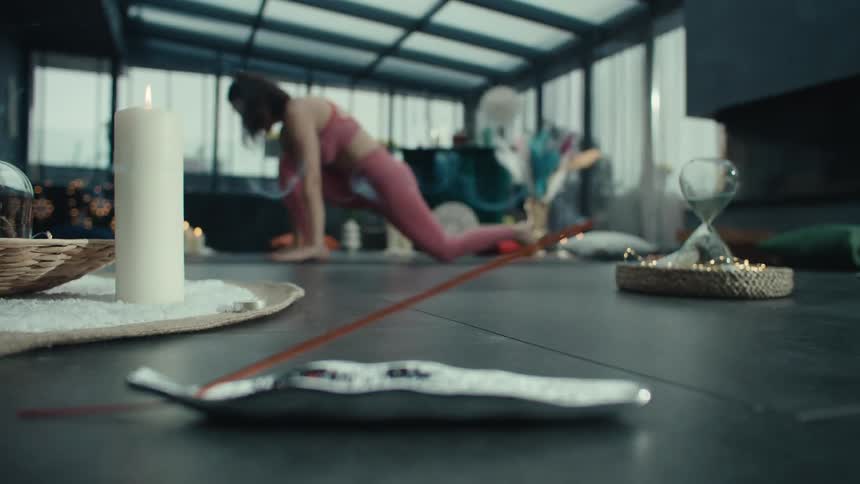 A woman in pink activewear performs yoga transitions and stretches in a glass walled studio while incense burns in the foreground next to a lit candle and an hourglass