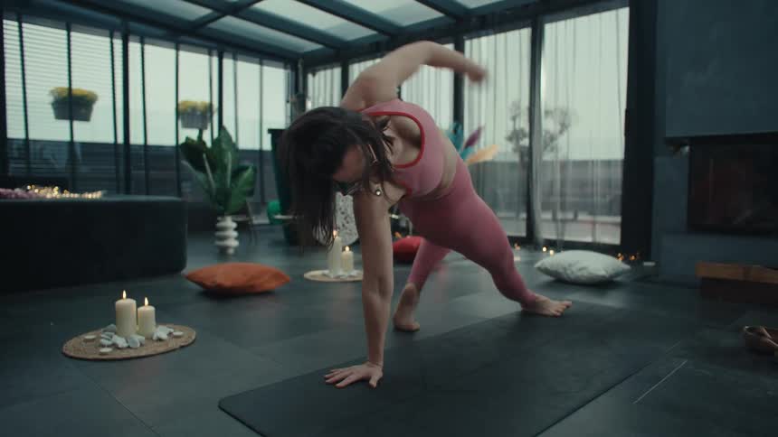 A woman in pink activewear performs a yoga sequence in a glass walled room moving from a wild thing pose into a plank and finishing in an upward facing dog surrounded by candles and decor