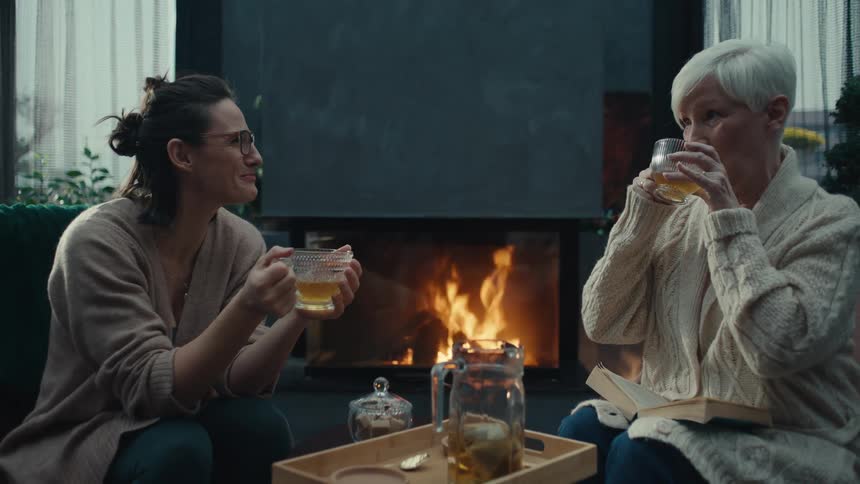 Two women sit comfortably by a roaring fireplace sharing tea and conversation in a cozy dimly lit roomthe younger woman wears glasses and a cardigan while listening intently to the older woman who rests a book on her lap as they enjoy their warm dri
