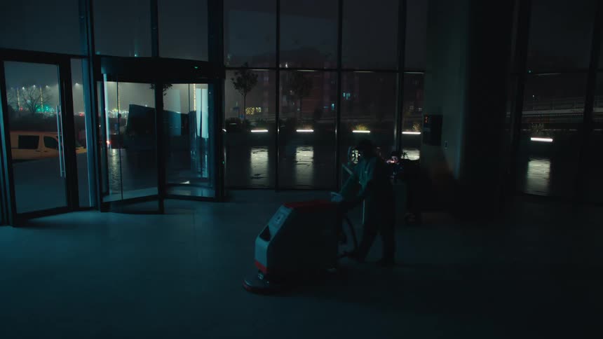 Janitor pushing his cleaning machine across the floor inside a building as traffic on the street is seen through the windows
