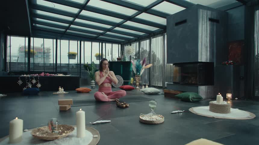 A woman in pink athletic wear sits crosslegged on a yoga mat in a modern sunlit room moving her arms in a meditative sequence