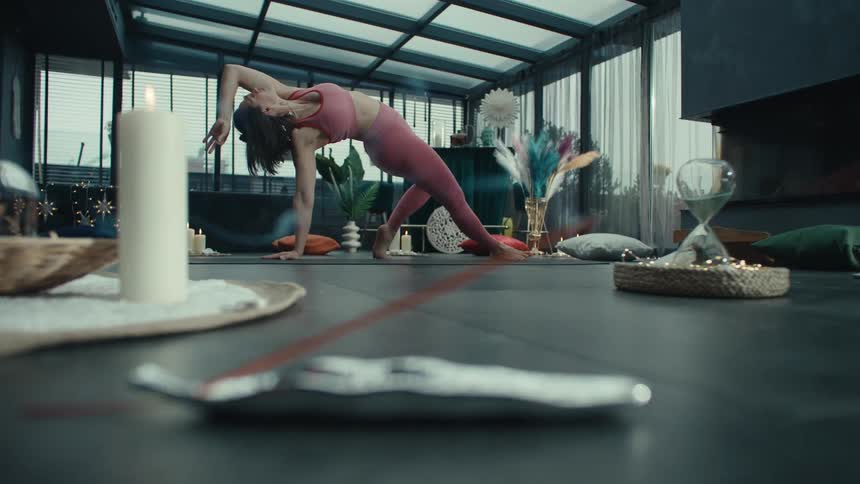 In the foreground a thin stick of incense slowly burns in a silver holder alongside a white candlein the blurred background a woman in pink athletic wear performs a side plank yoga pose within a modern glassroofed room