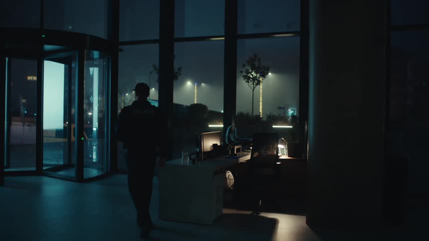 A bearded security guard in a black uniform pours a hot drink into a glass mug and walks through a dark office lobby toward a revolving door at night