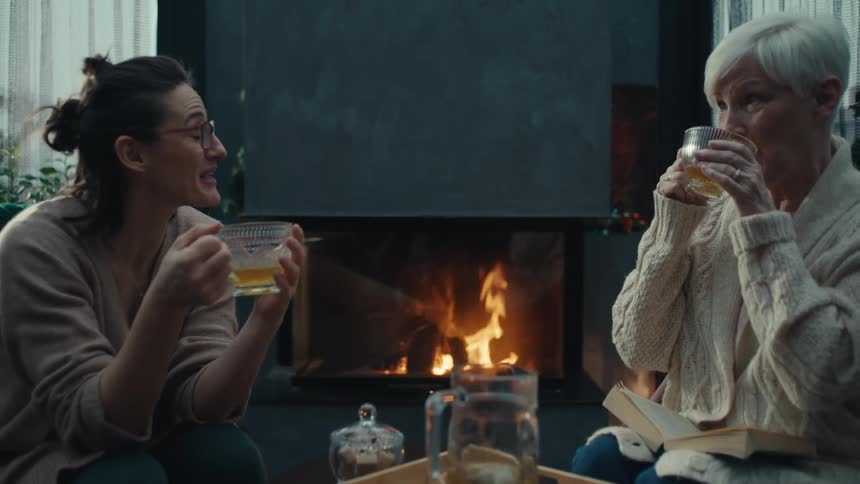 Two women sitting at a table drinking tea and talking as a fire burns in the fireplace