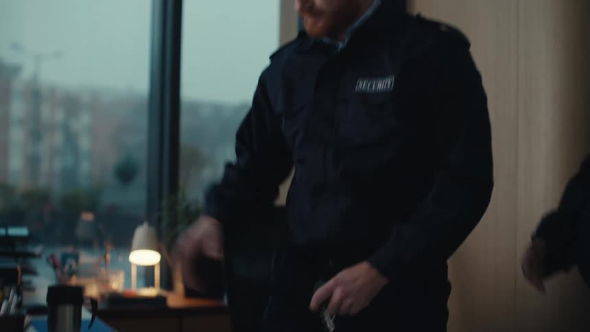 A bearded security guard sleeping at a desk is startled awake by a coworker who then takes over the workstation and begins using the computer