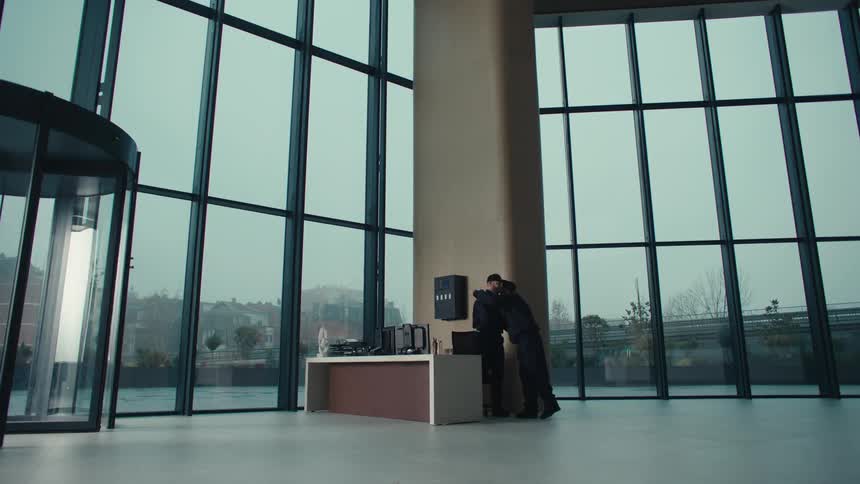 A security guard finishing his shift is replaced by another and the two share a quick hug in the lobby of an office building at midday while employees stream inside