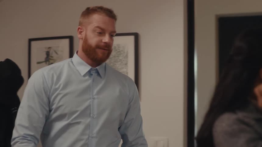 A man with a red beard enters a home takes off his jacket and black cap and hangs them on a coat rackhe then approaches a woman who is adjusting her hair in a mirror and gently touches her shoulder before she continues grooming herself