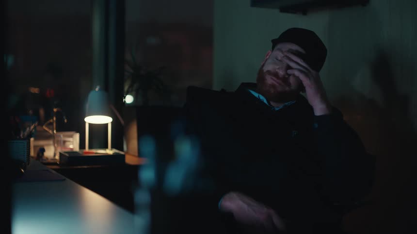 A male security guard wearing a black uniform and cap sits at a dark desk in front of a computer screen then he yawns rubs his face with his hand and eventually leans his head back against the chair to fall asleep during his night shift