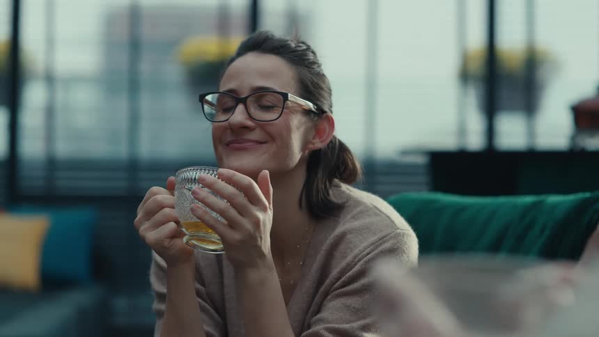 A woman wearing glasses sits comfortably on a couch while holding a warm glass of tea she takes a slow sip with her eyes closed then smiles and talks warmly to someone offcamera