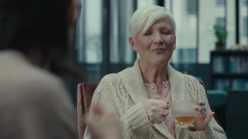 An elegant older woman with short white hair sits in a modern interior smiling warmly and engaging in conversation while holding a clear teacupas she chats with her companion she takes a gentle sip of her tea and continues to listen with a pleasant