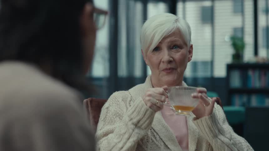 An elderly woman with short white hair smiles and sips tea while listening to a companionshe wears a creamcolored knit cardigan and engages in a cheerful relaxed conversation in a brightly lit indoor space