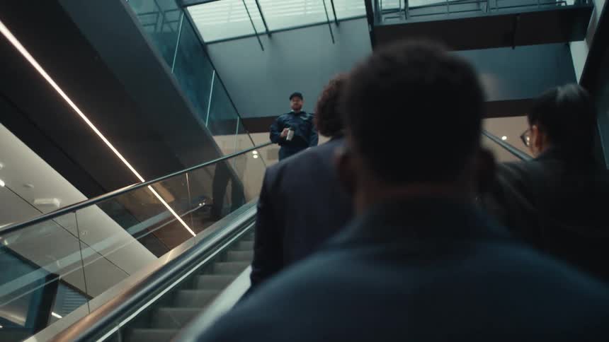 Young adult black man on an office space walking and going up mechanical stairs with other people and then greeting a smiling man wearing a baseball cap on the stairs to the side indoors during day time