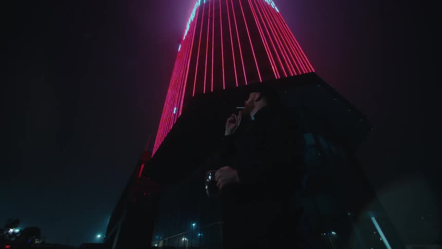 A bearded security guard in a black uniform stands outside at night holding a mug and exhaling a cloud of vapor while looking up at a tall building illuminated with vertical red lights