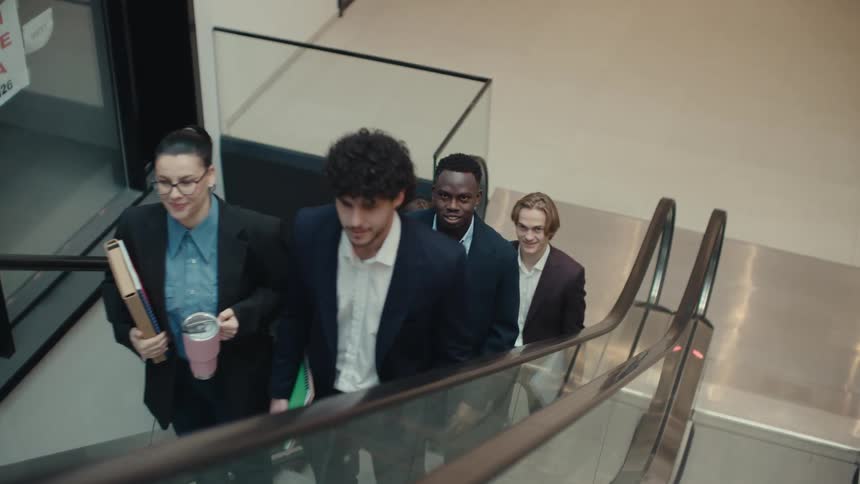 A group of employees walks toward an escalator inside an office building on a busy workday then rides it up to the next floor