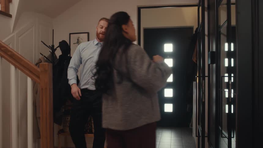 A man in a light blue shirt enters a home takes off his jacket and hat and greets a woman who is adjusting her hair in a mirror
