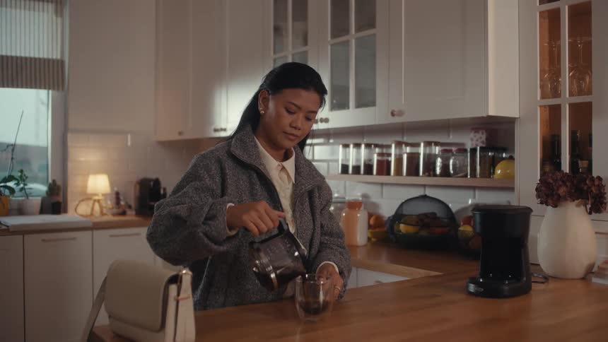 A young woman wearing a winter jacket walks into the kitchen picks up a glass cup pours coffee into it and takes a sip