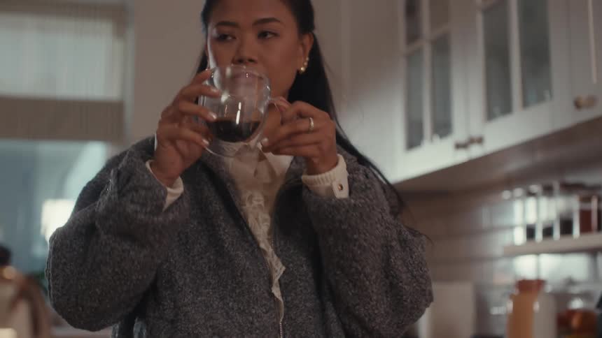 Woman stands in the kitchen pouring coffee into a glass cup and taking a drink