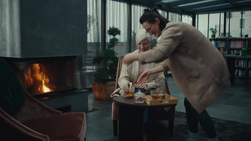 A woman brings a tray with tea and sits down with another woman by the fire