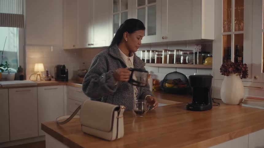 Woman walks into the kitchen and takes a glass mug out of the cupboard then pours a cup of coffee and takes a drink before leaving