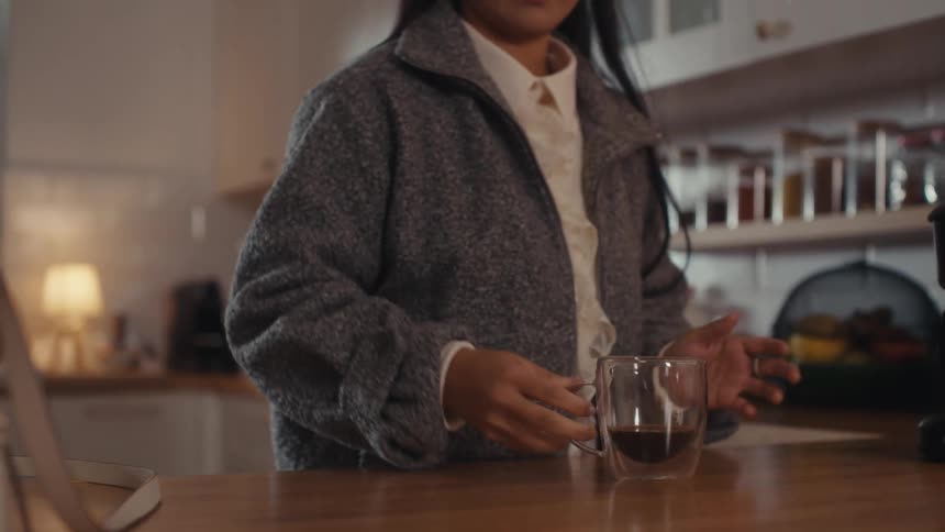 Woman standing in the kitchen pours coffee into a glass cup and takes a drink