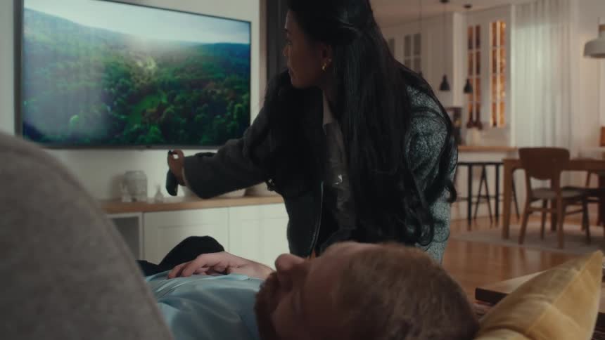 Woman turning off the tv and adjusting a blanket over a man lying on a sofa in a domestic setting