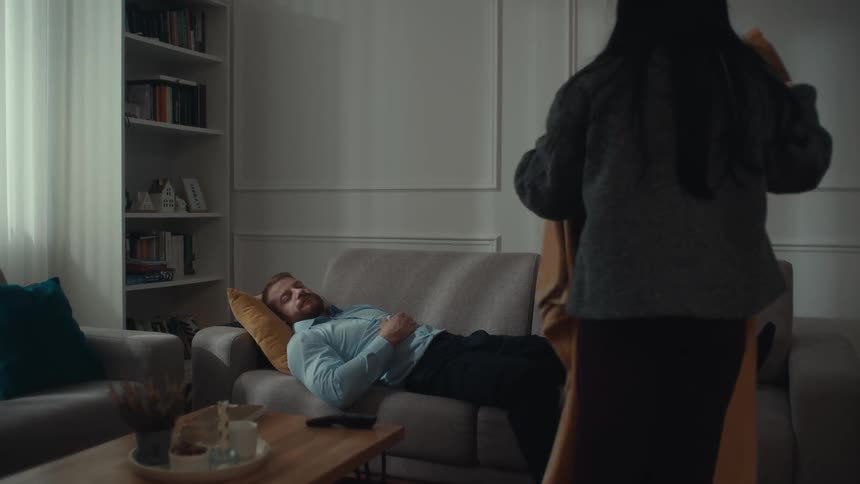 A young woman gently covers a sleeping man with a yellow blanket as he rests on a grey sofa in a living room
