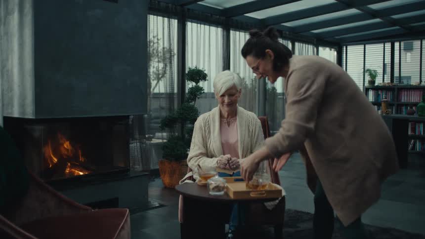 Woman serves tea and sits down with an elderly woman and they both drink