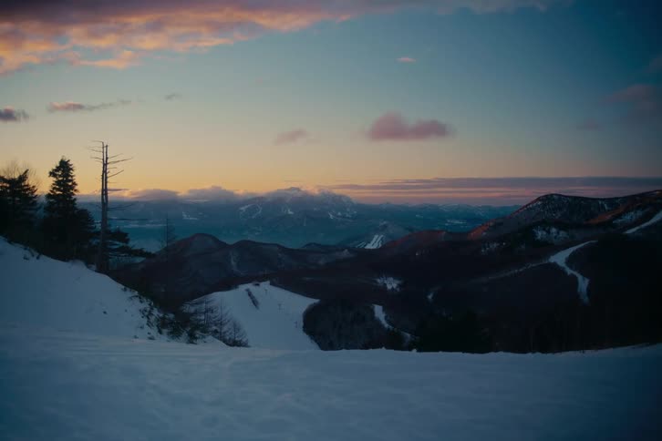 A serene snowcovered mountain landscape sits under a soft twilight sky filled with hues of orange and purplebelow the rolling peaks a quiet valley reveals winding ski runs and a distant body of water reflecting the fading light