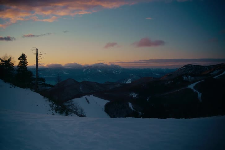 A wide alpine landscape reveals snow covered mountains and ski slopes under a vibrant twilight sky