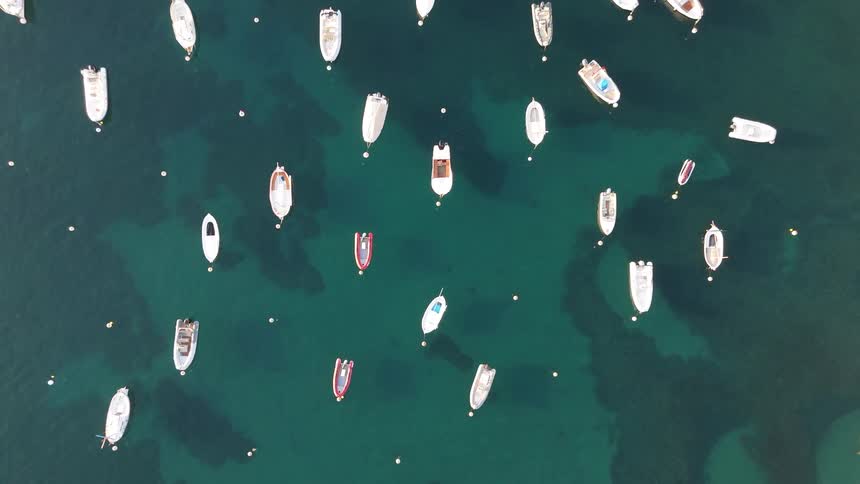 A top down aerial view shows many small white and red boats floating on clear teal water