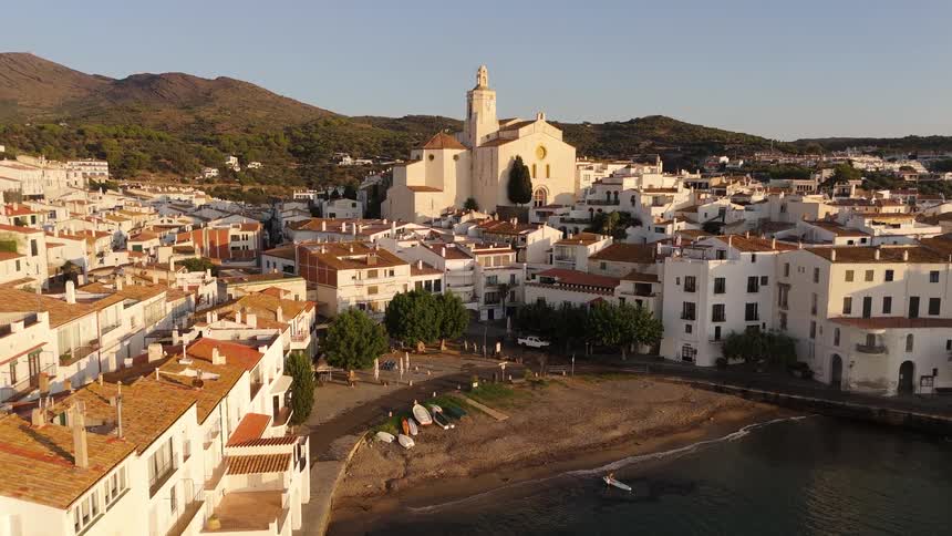An aerial view shows a coastal town with white buildings and red tile roofs situated along a small beach during sunset