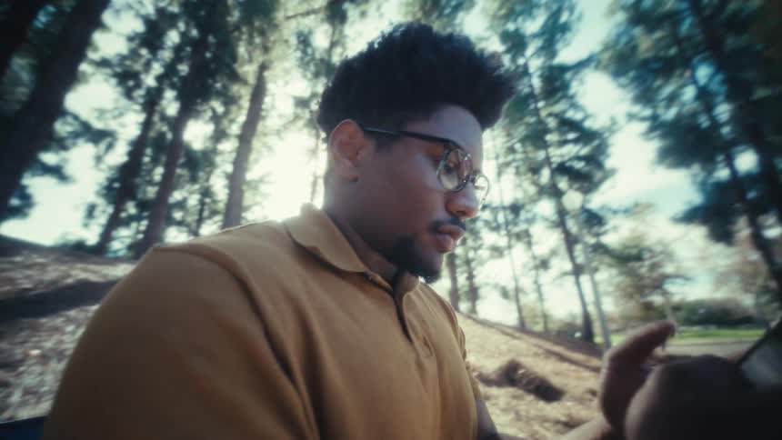 A man with dark curly hair and glasses sits on a park bench while focused on his smartphonethe camera moves in a low sweeping arc around him capturing the sunlight filtering through the tall trees in the background