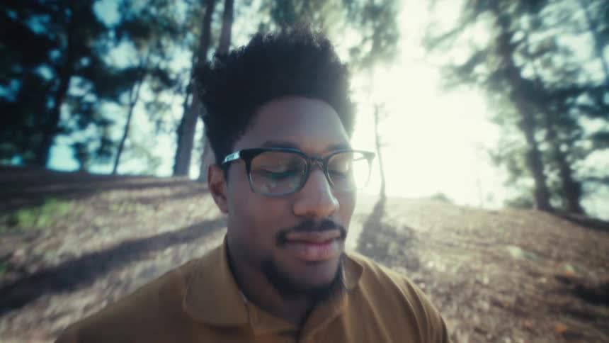 A man wearing glasses sits on a park bench and texts on his phone as sunlight filters through the trees behind him