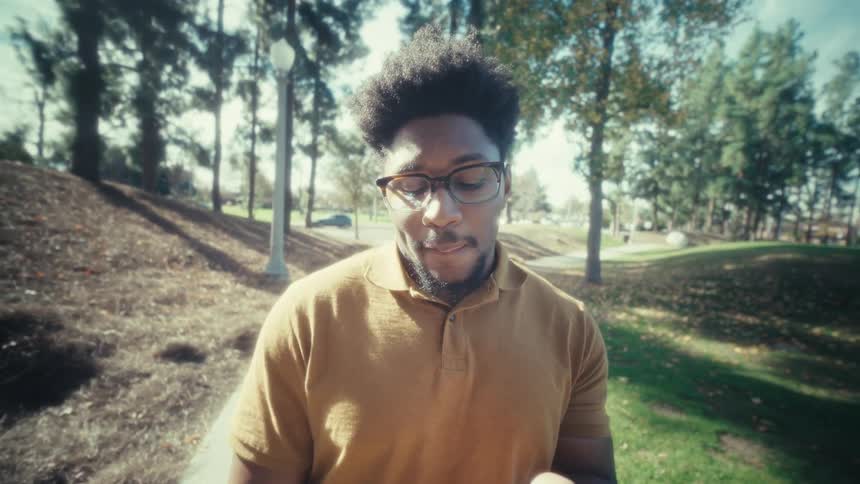 A man typing on a smartphone while sitting in a public park in bright daylight