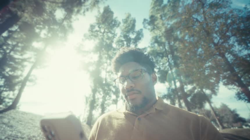 A man with glasses and a beard stands in a sunlit park with tall trees and looks at his phone