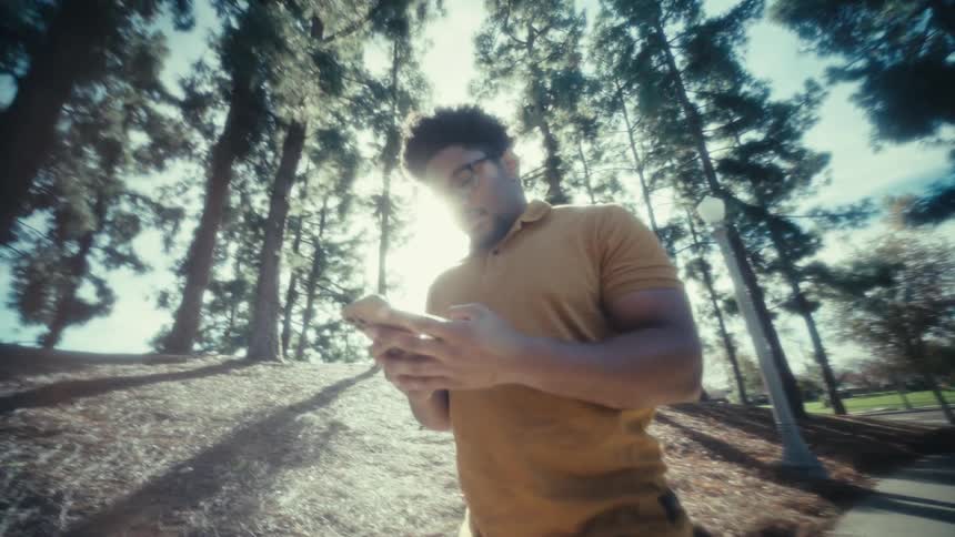 Young adult black man walking on a park next to a road and picking up his phone to text while smiling outdoors during day time