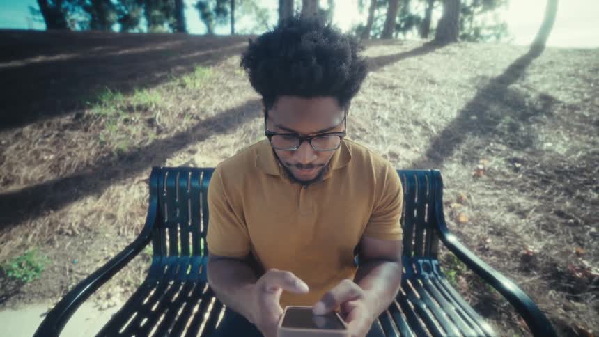 A man sitting on a park bench while typing on a smartphone in bright daylight at a public park